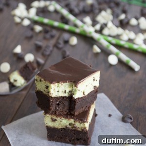 Three stacked chocolate brownies with a creamy mint layer in the middle are placed on a piece of parchment paper. White and dark chocolate chips and green straws are scattered on a wooden surface in the background.
