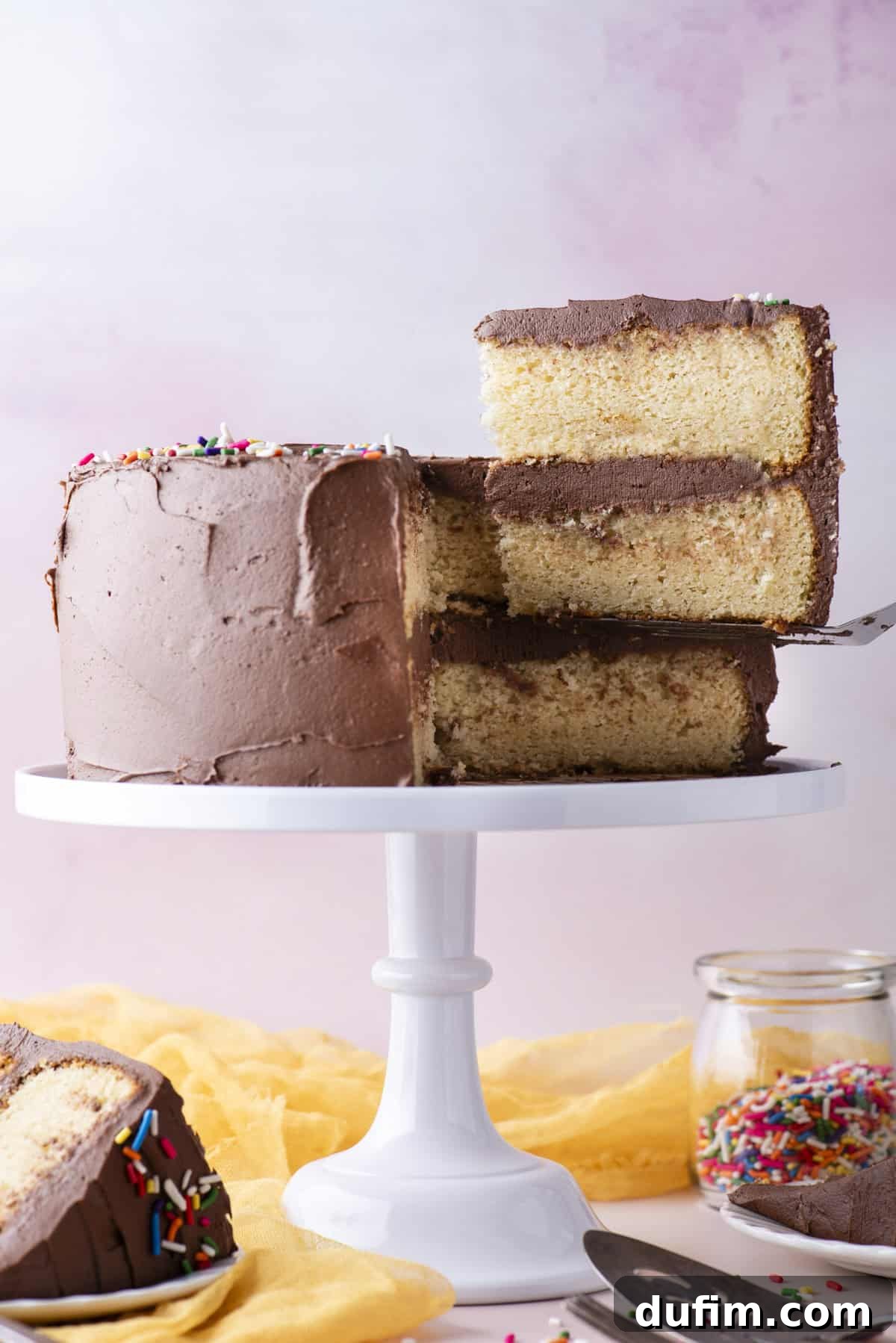 A delectable slice of two-layer yellow gluten-free cake with rich chocolate frosting, being gracefully lifted from a white cake stand. A single slice rests on a white plate, accompanied by a fork, a metal spatula, a cheerful yellow cloth, and a clear glass jar brimming with rainbow sprinkles below.