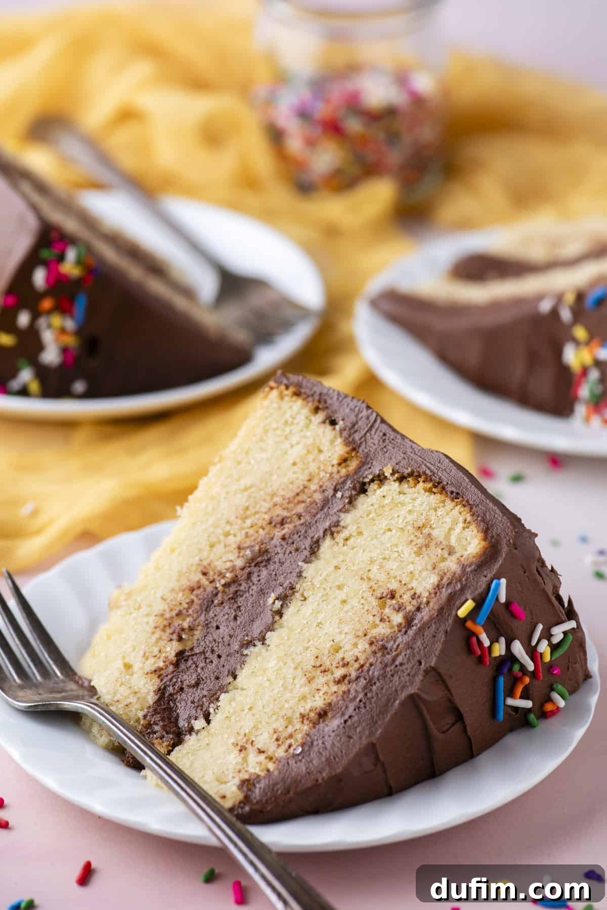 A delicious slice of two-layer vanilla cake, generously covered in chocolate frosting and rainbow sprinkles, lies invitingly on its side on a small white plate, accompanied by a fork. The setting is a light pink surface, with additional cake slices on plates and a jar of sprinkles subtly blurred in the background.