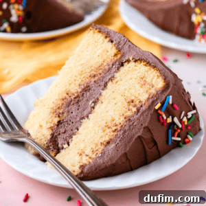 A slice of two-layer vanilla cake with chocolate frosting and rainbow sprinkles laying on its side on a small white plate with a fork, sitting on a light pink surface with more slices on plates in the background