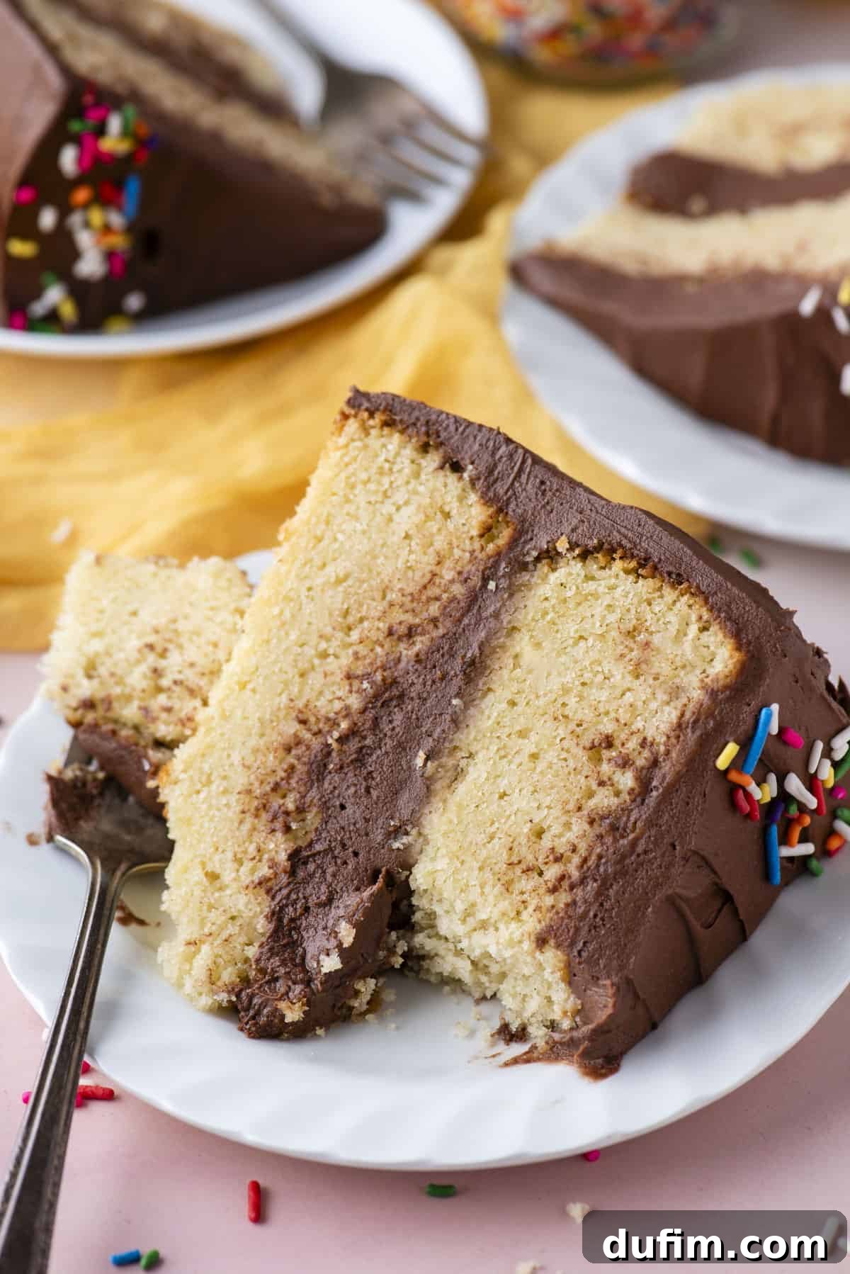 A beautifully presented slice of two-layer yellow cake, adorned with rich chocolate frosting, rests elegantly on a white plate. A fork, holding a single bite of cake, leans against the slice. The setting features a light pink surface, with additional frosted cake slices on plates softly blurred in the background, and charming rainbow sprinkles scattered around.