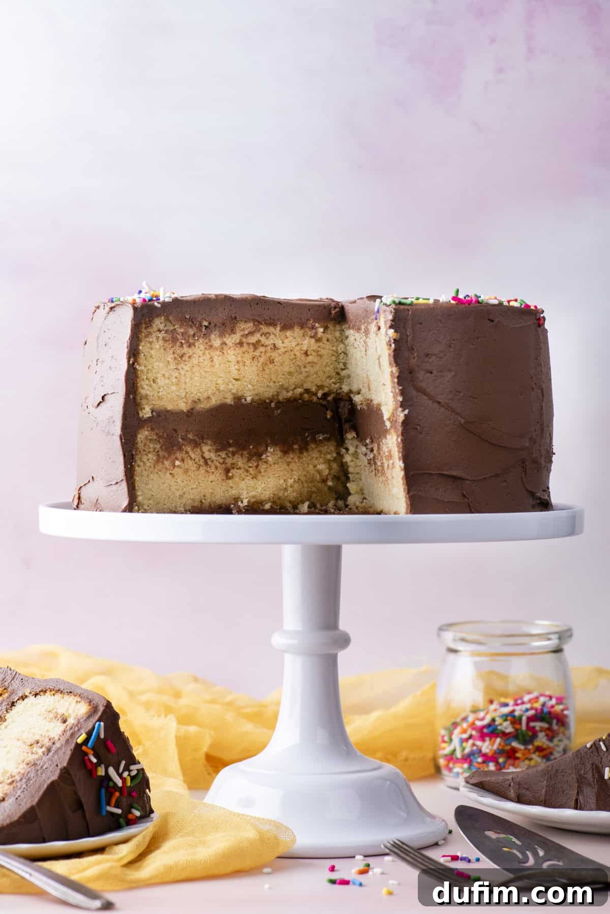 A beautifully decorated two-layer gluten-free cake with smooth chocolate frosting and festive rainbow sprinkles generously scattered on top. It sits proudly on a white cake stand, accompanied by a cheerful yellow cloth, two plates holding slices of cake, a serving fork, a metal serving spatula, and a clear glass jar filled with extra rainbow sprinkles below.