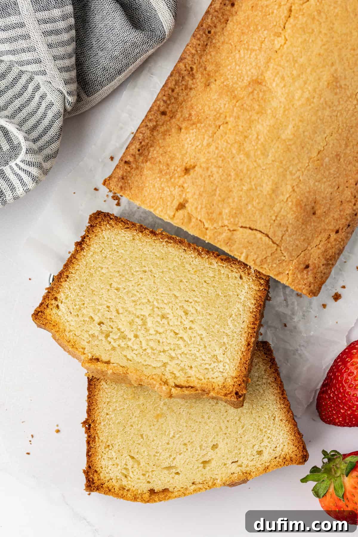 Overhead view of a golden-brown pound cake with two neat slices cut, adorned with fresh strawberries, beside a stylish grey and white striped kitchen towel. A perfect presentation of a classic dessert.