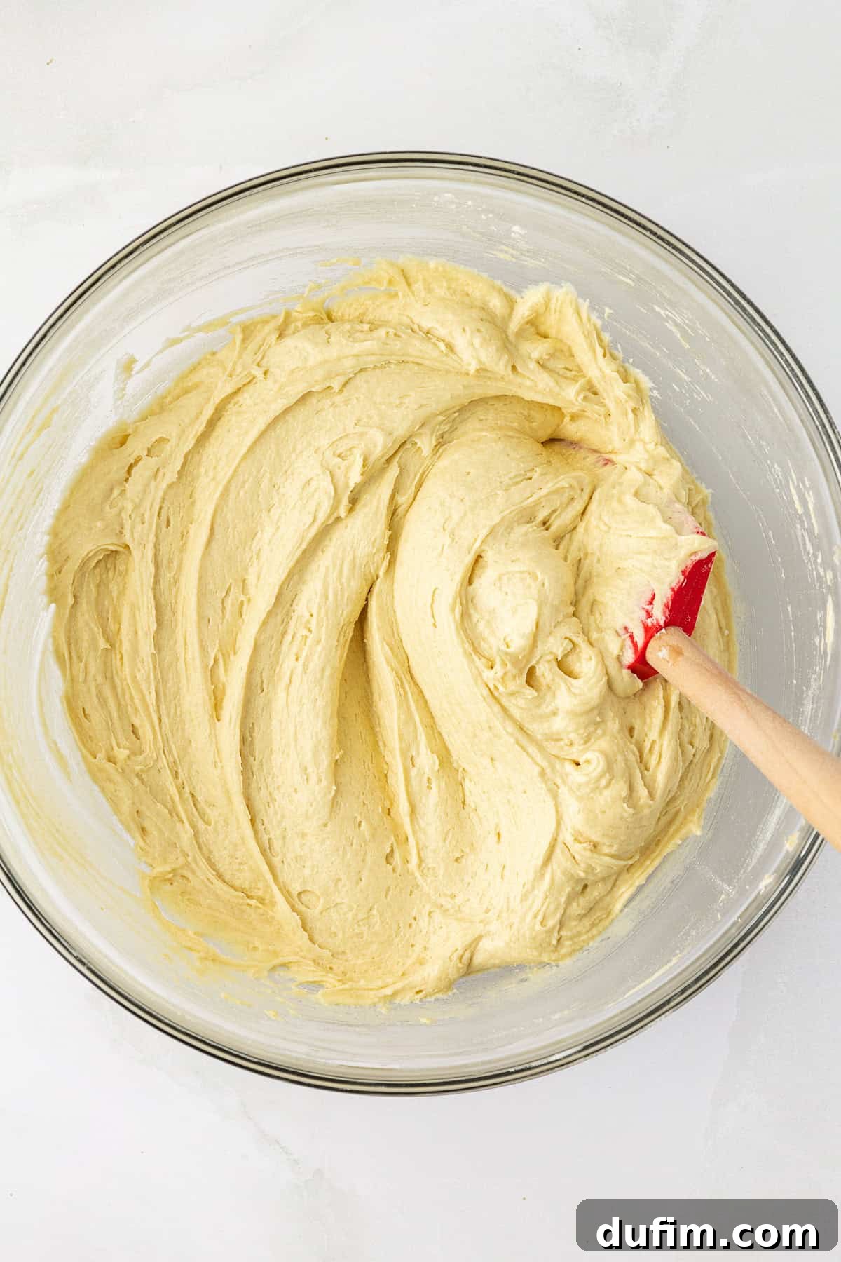 Thick, smooth pound cake batter filling a clear glass bowl, with a red and wooden spatula resting inside, demonstrating the perfect consistency before transfer to the baking pan.