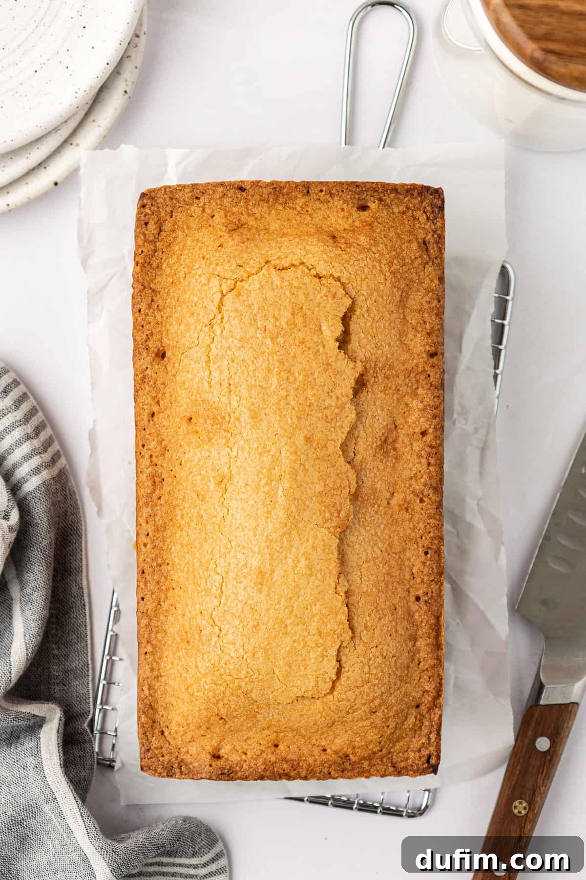 A perfectly baked, whole pound cake resting on parchment paper on a wire rack, flanked by a soft grey kitchen towel and a stack of white plates to its left, with a sharp knife positioned to its right, inviting a slice.