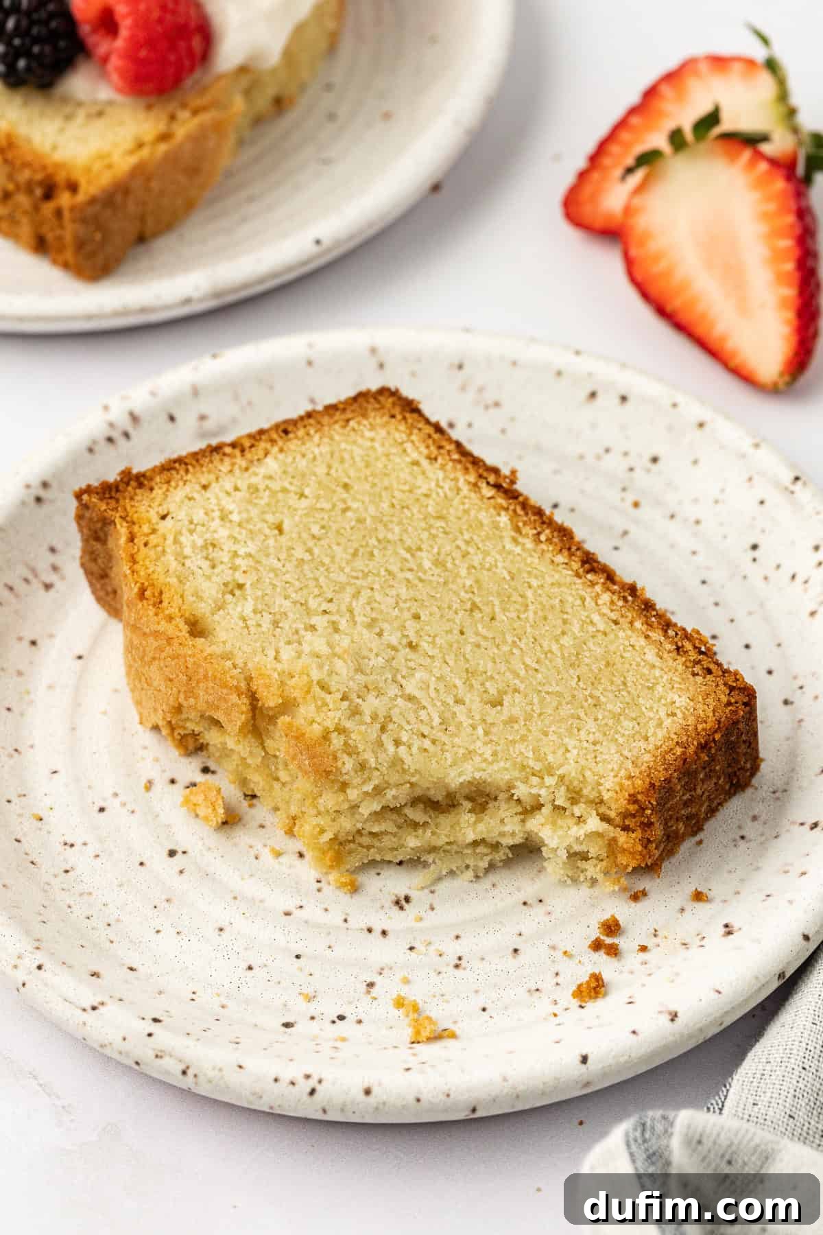 A charming slice of pound cake on a white speckled plate, with a single bite missing, indicating its deliciousness. Nearby, a fork leans casually against the plate, and in the background, another slice of pound cake adorned with whipped cream and berries, alongside a bowl of fresh strawberries, completes this inviting dessert scene.