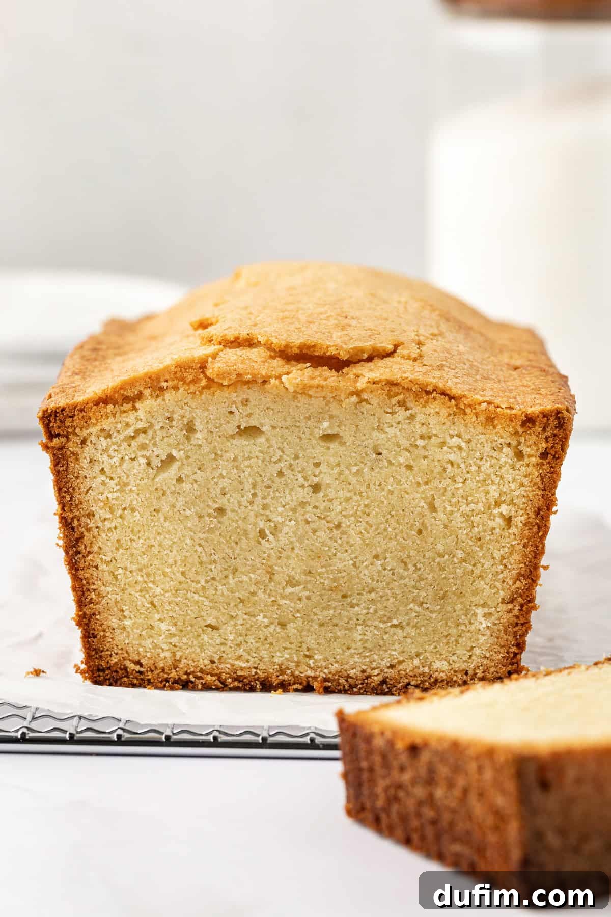 A beautifully baked pound cake on parchment paper, resting on a wire rack. One perfect slice is carefully cut and placed beside the main cake, showcasing its inviting light yellow interior and gorgeously golden brown crust. A true testament to a successful homemade pound cake.