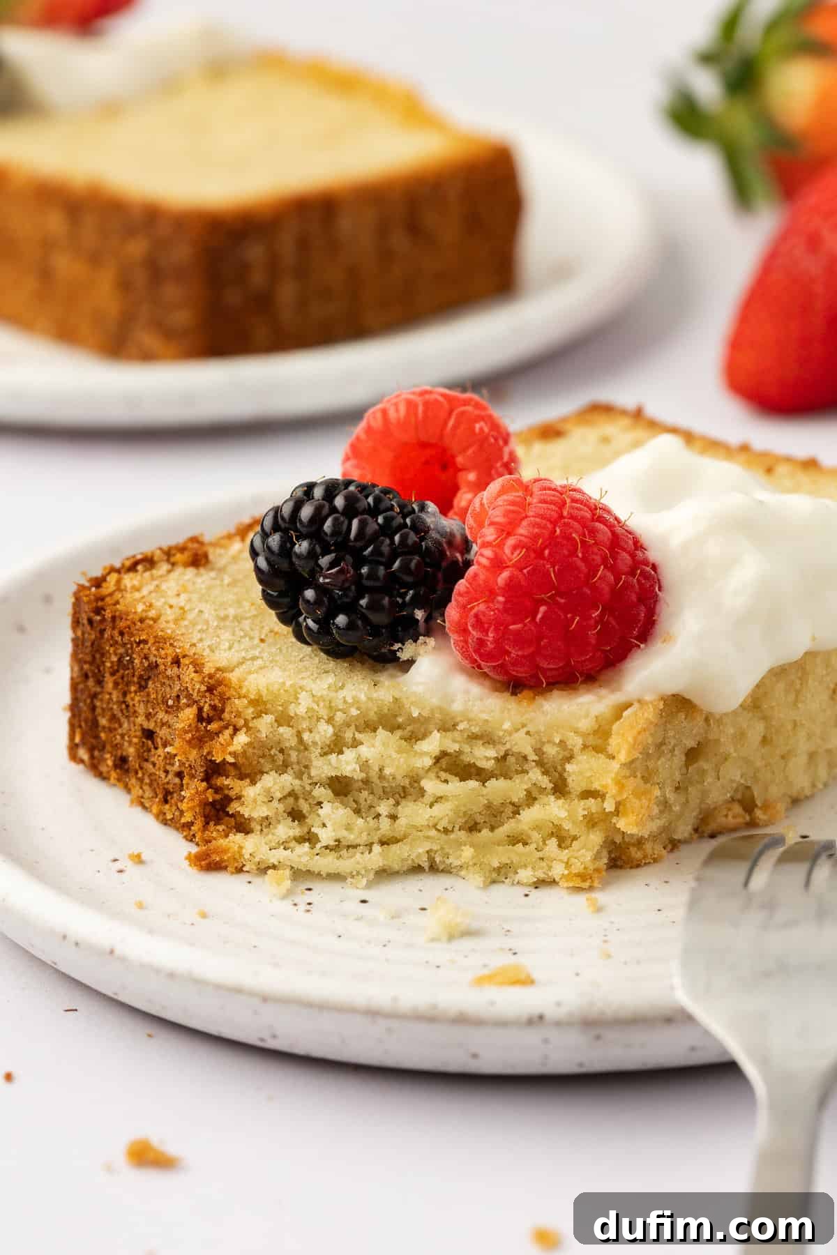 A mouthwatering slice of golden pound cake, elegantly topped with fluffy whipped cream and a colorful array of fresh berries. A single bite is taken, leaving a tempting indentation. In the soft-focused background, a fork rests on the plate, another berry-topped slice of cake, and a bowl of fresh strawberries complete this delicious scene.