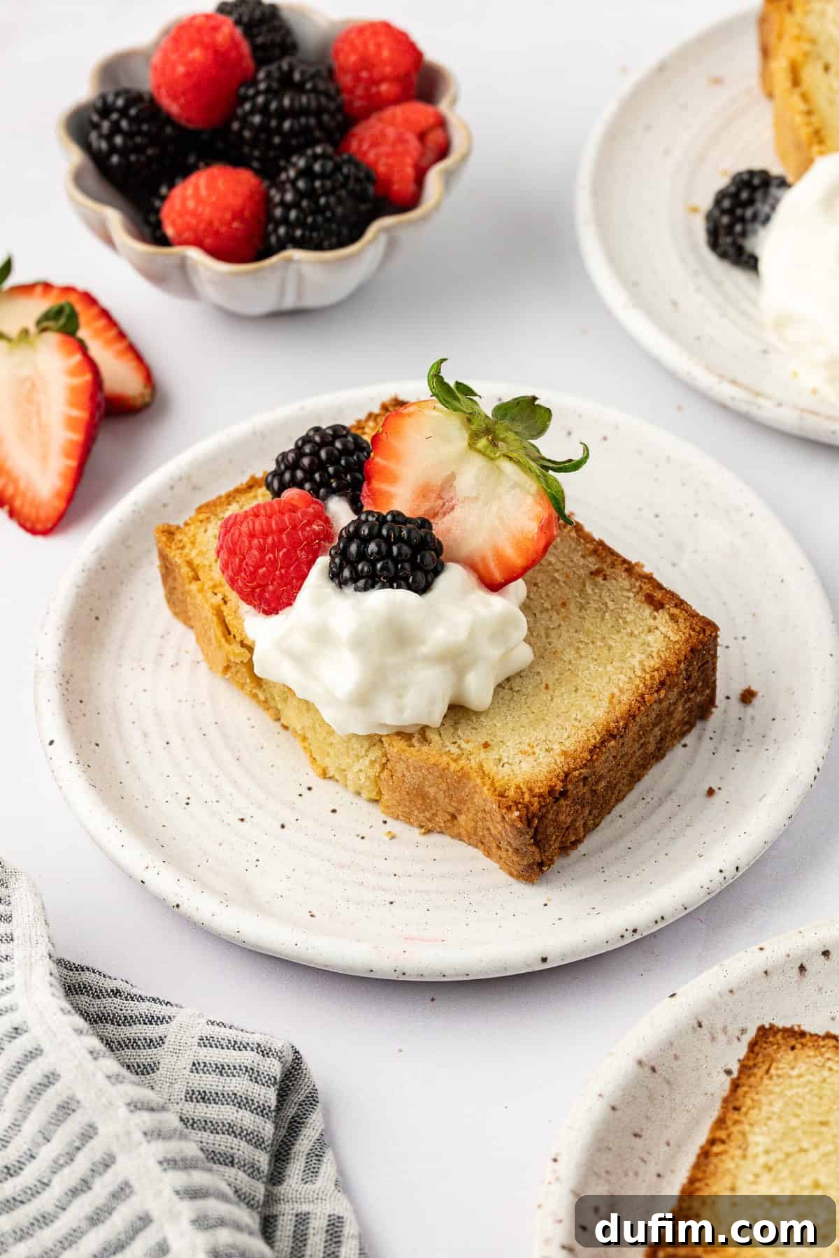 A delectable slice of pound cake on a pristine white plate, generously topped with airy whipped cream and a vibrant medley of fresh berries. Surrounding it are more plates with slices, a bowl overflowing with fresh berries, and neatly sliced strawberries, all inviting a delightful indulgence.