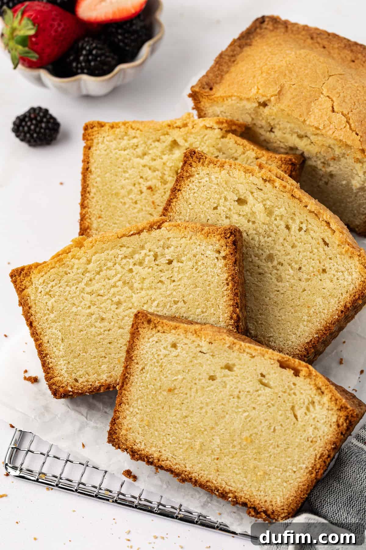 Perfectly baked slices of golden pound cake artfully leaned against each other on a wire cooling rack, resting on parchment paper. A generous bowl of vibrant fresh berries sits invitingly beside it, promising a delightful pairing.