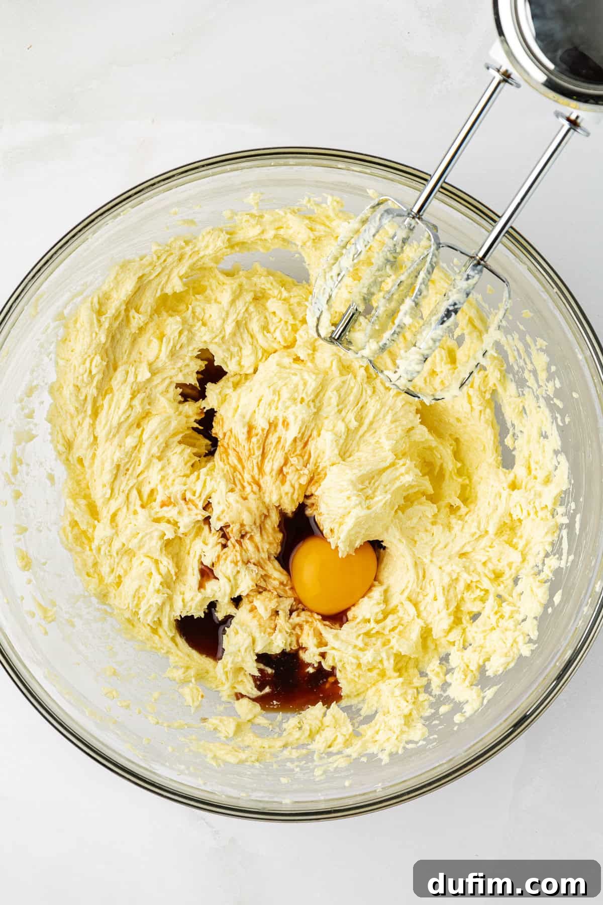 The rich mixture of creamed butter and sugar in a clear glass bowl, with whole eggs and fragrant vanilla extract carefully added on top, awaiting integration into the pound cake batter, an electric mixer resting on the side.
