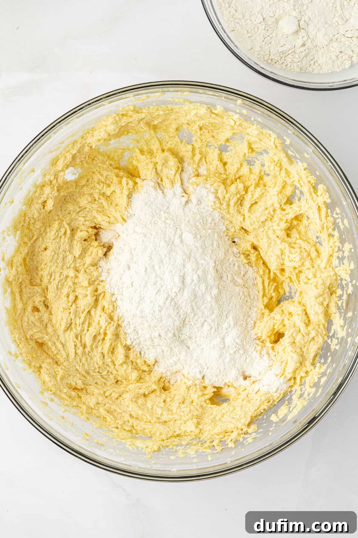 The combined wet ingredients in a clear glass bowl, topped with a mound of the dry flour mixture, and a separate small bowl holding remaining dry ingredients. This setup illustrates the gradual mixing process for pound cake.