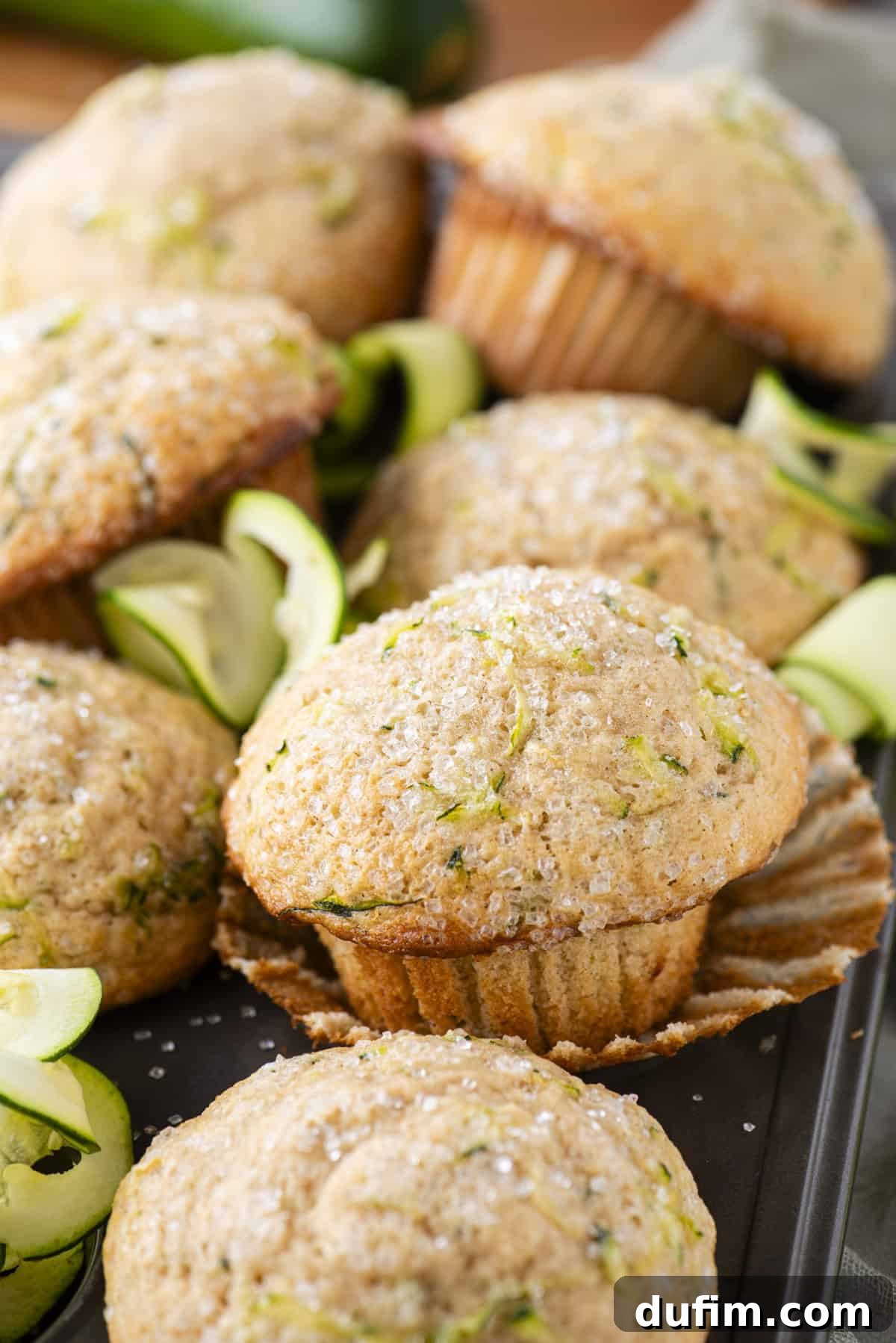 A delightful stack of golden-brown zucchini muffins, some with their liners partially peeled, showcasing the tender crumb and subtle green flecks of zucchini. Thin, delicate zucchini slices are artfully arranged around them, creating an inviting culinary scene.