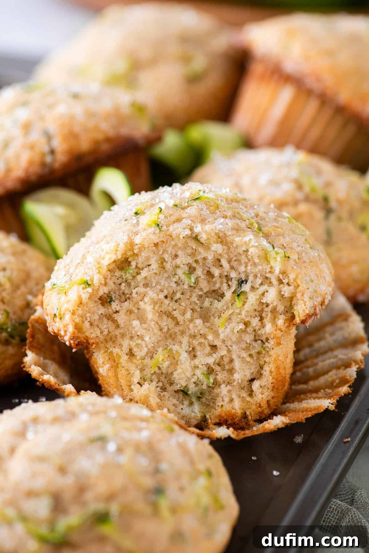 A mound of freshly baked zucchini muffins rests on a muffin pan, with one in the center gently peeled from its liner, revealing a tender crumb and a missing bite, tempting the viewer to try.