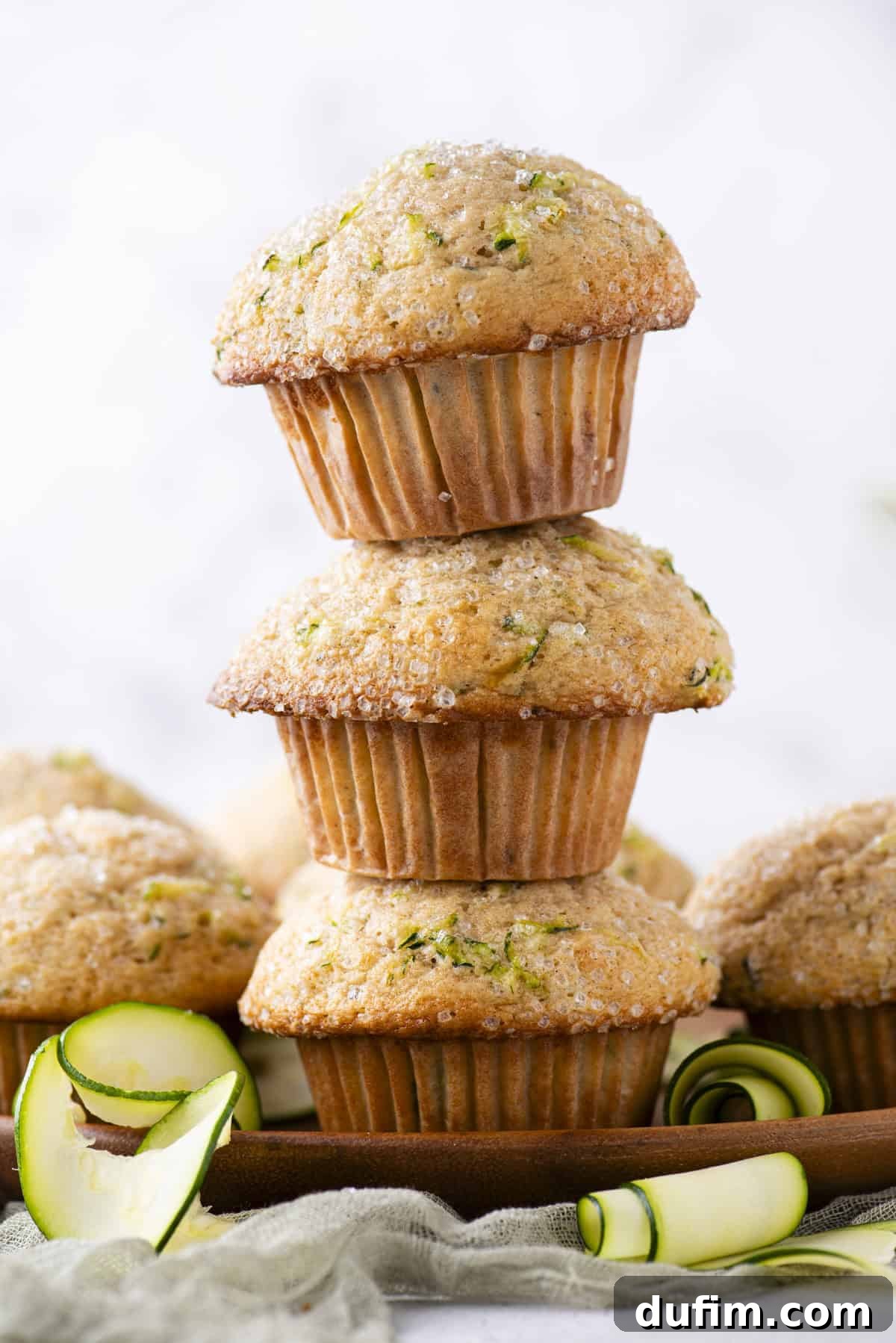 A charming pyramid of three fluffy zucchini muffins stacked on a rustic wooden plate, surrounded by more freshly baked muffins and delicate curls of zucchini peel, inviting you to indulge.