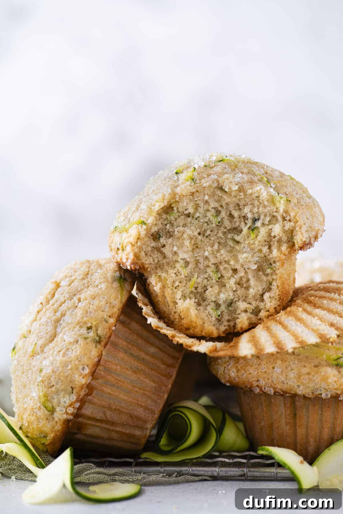 A delectable zucchini muffin, with its liner removed and a bite taken, sits atop a wire cooling rack. More muffins blur softly in the background, amidst scattered delicate zucchini slices, highlighting the fresh, homemade appeal.