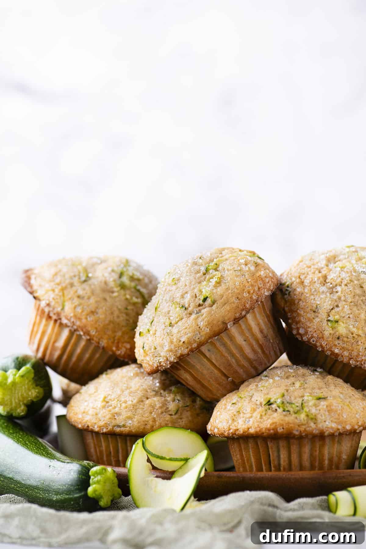 A generous pile of freshly baked zucchini muffins sits on a wooden plate, surrounded by fresh, whole zucchinis and delicate slices, emphasizing the natural, wholesome ingredients.
