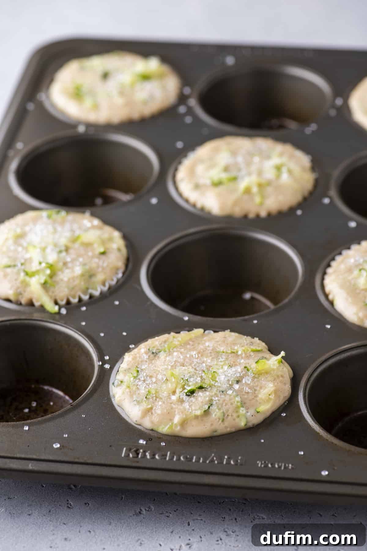 A muffin pan with every other well filled generously with thick zucchini muffin batter, showing the strategic spacing for optimal rise.