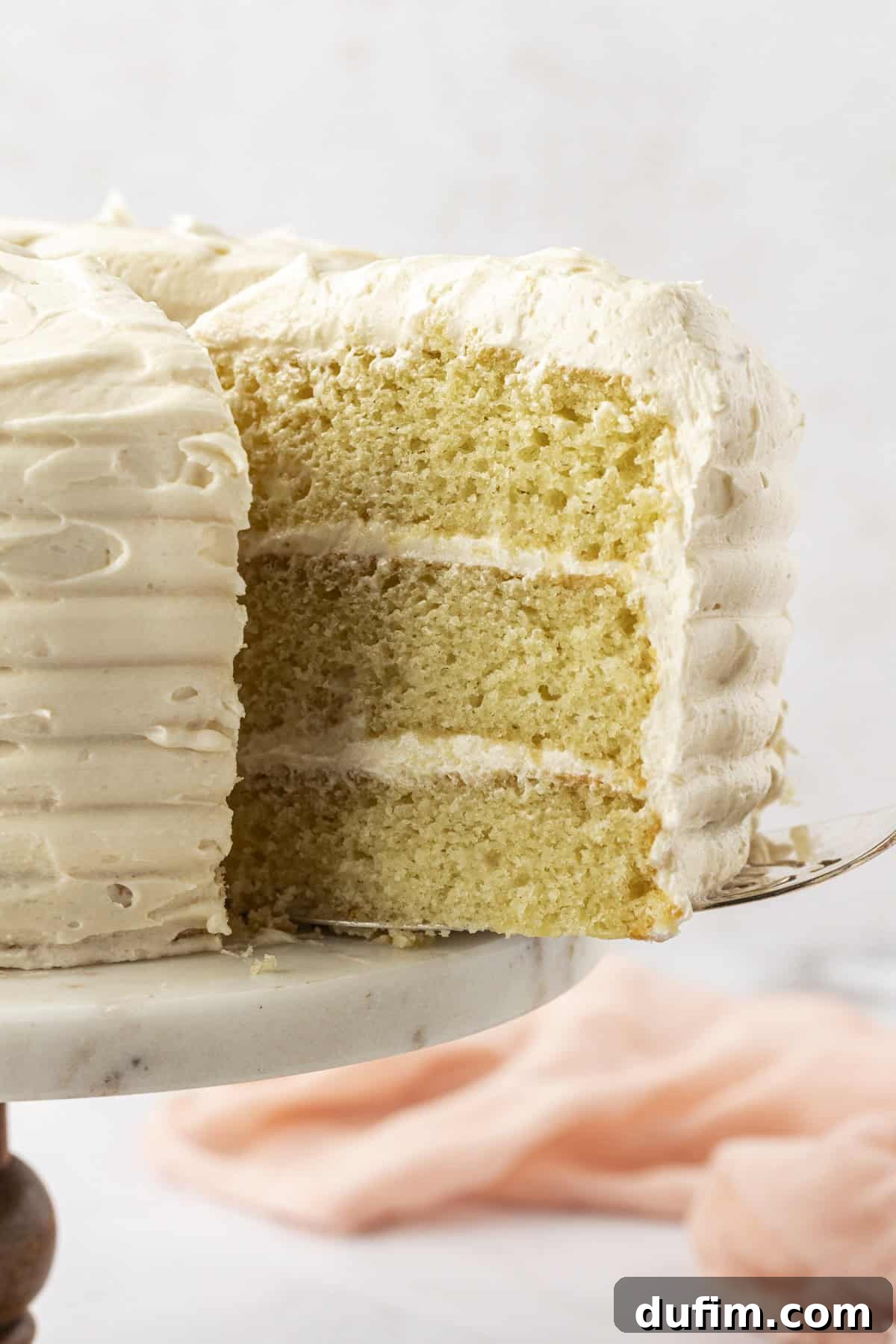 A beautifully layered three-tier vanilla cake with creamy vanilla frosting, a slice being lifted from the cake stand on a pink cloth.