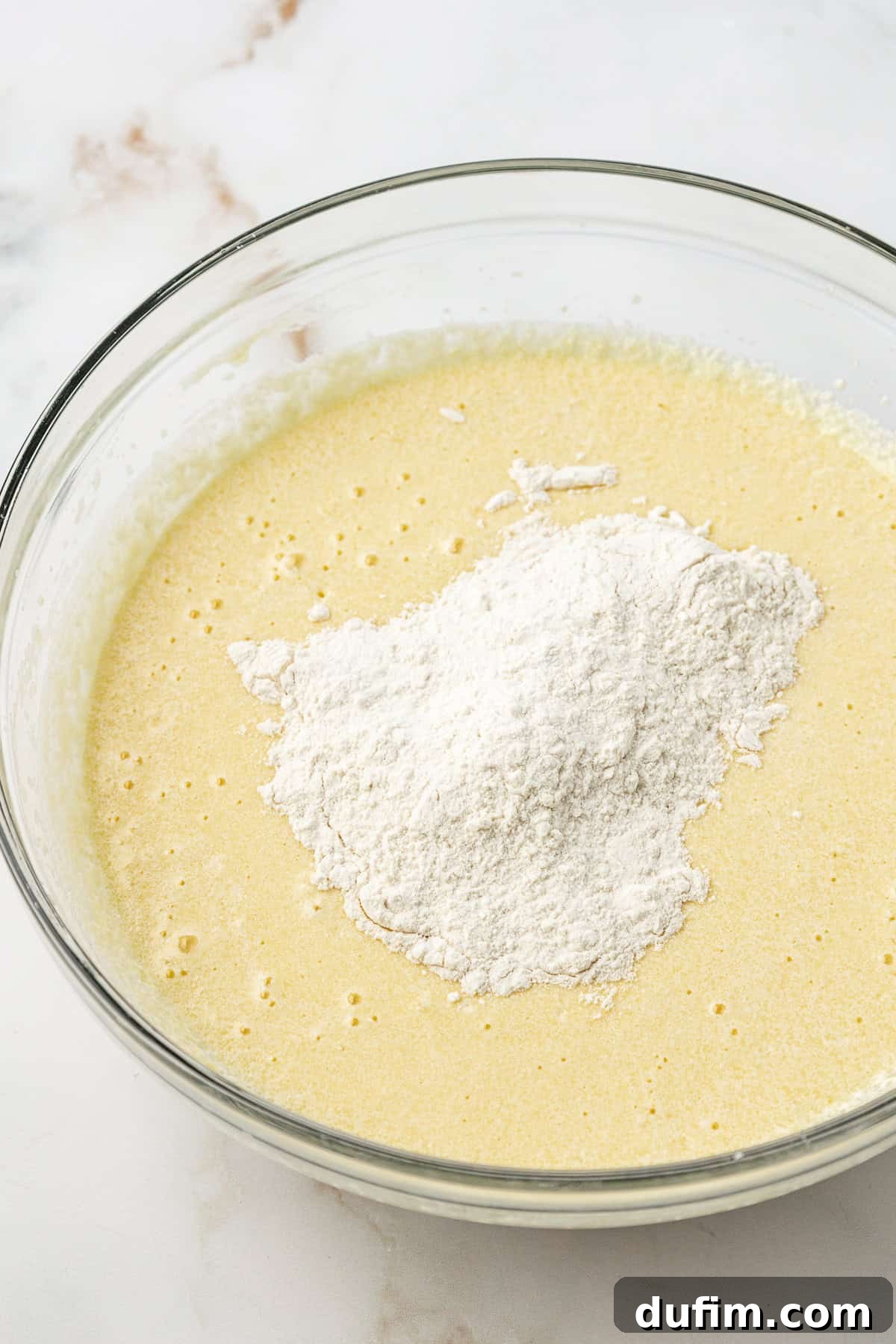 The wet ingredients for vanilla cake in a clear glass bowl, with the flour mixture gently added on top, ready to be combined.