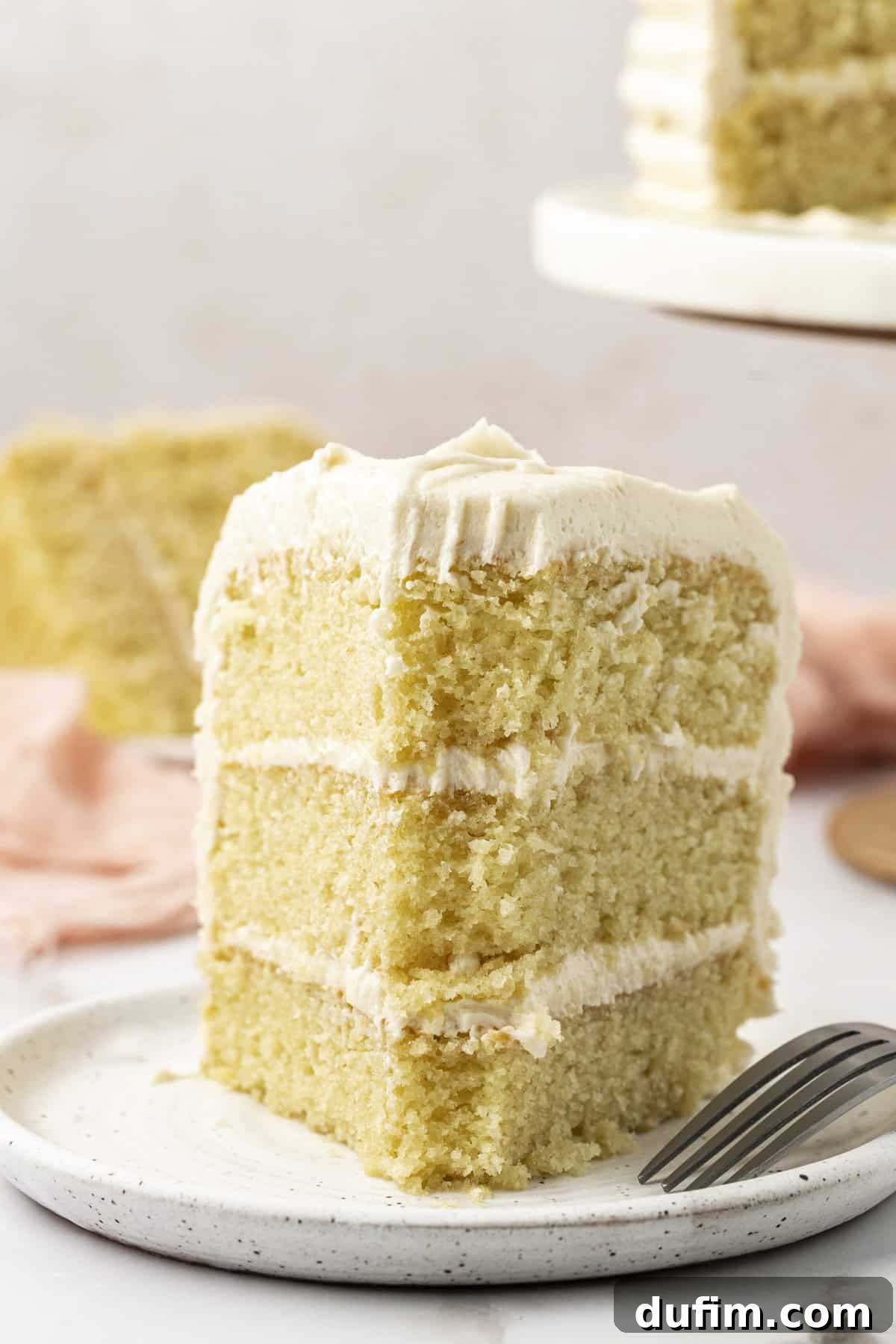 A slice of three-layer vanilla cake on a white speckled plate with a fork, showing the moist layers and creamy frosting. The main cake stand is visible in the background.