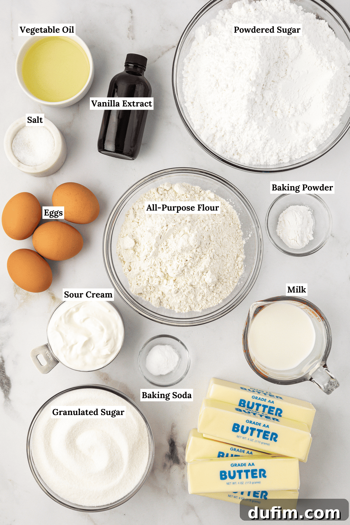 Overhead view of various vanilla cake ingredients laid out on a surface: vegetable oil, powdered sugar, vanilla extract, salt, eggs, all-purpose flour, baking powder, sour cream, baking soda, milk, granulated sugar, and butter.