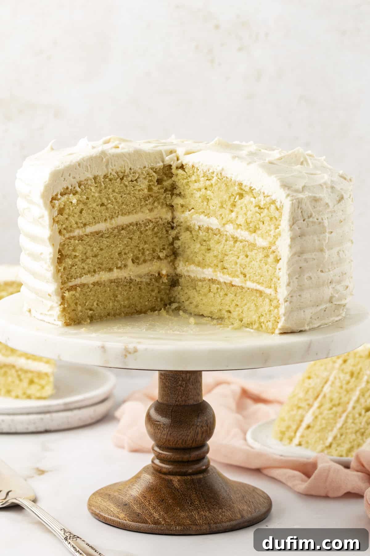 A beautiful three-layer vanilla cake on a cake stand with a large slice removed, revealing the moist interior. Two plates with cake slices are on a pink cloth below.