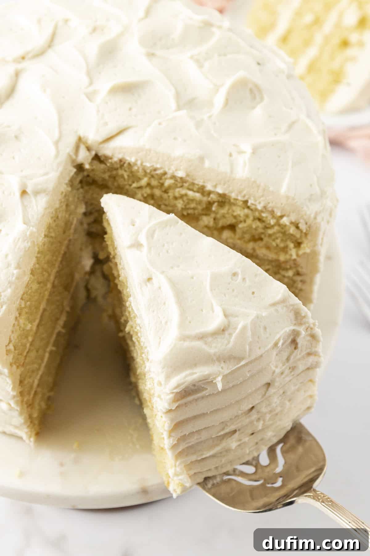 Overhead view of a slice of moist vanilla cake being carefully pulled away from the larger cake on a stand, highlighting its fluffy texture.