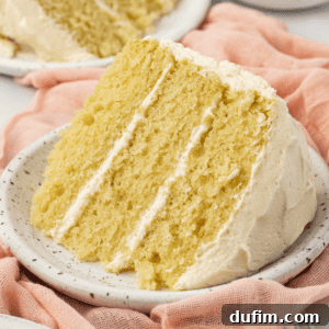 A slice of three layer vanilla cake on a white plate on top of a pink cloth with more cake slices on plates in the background