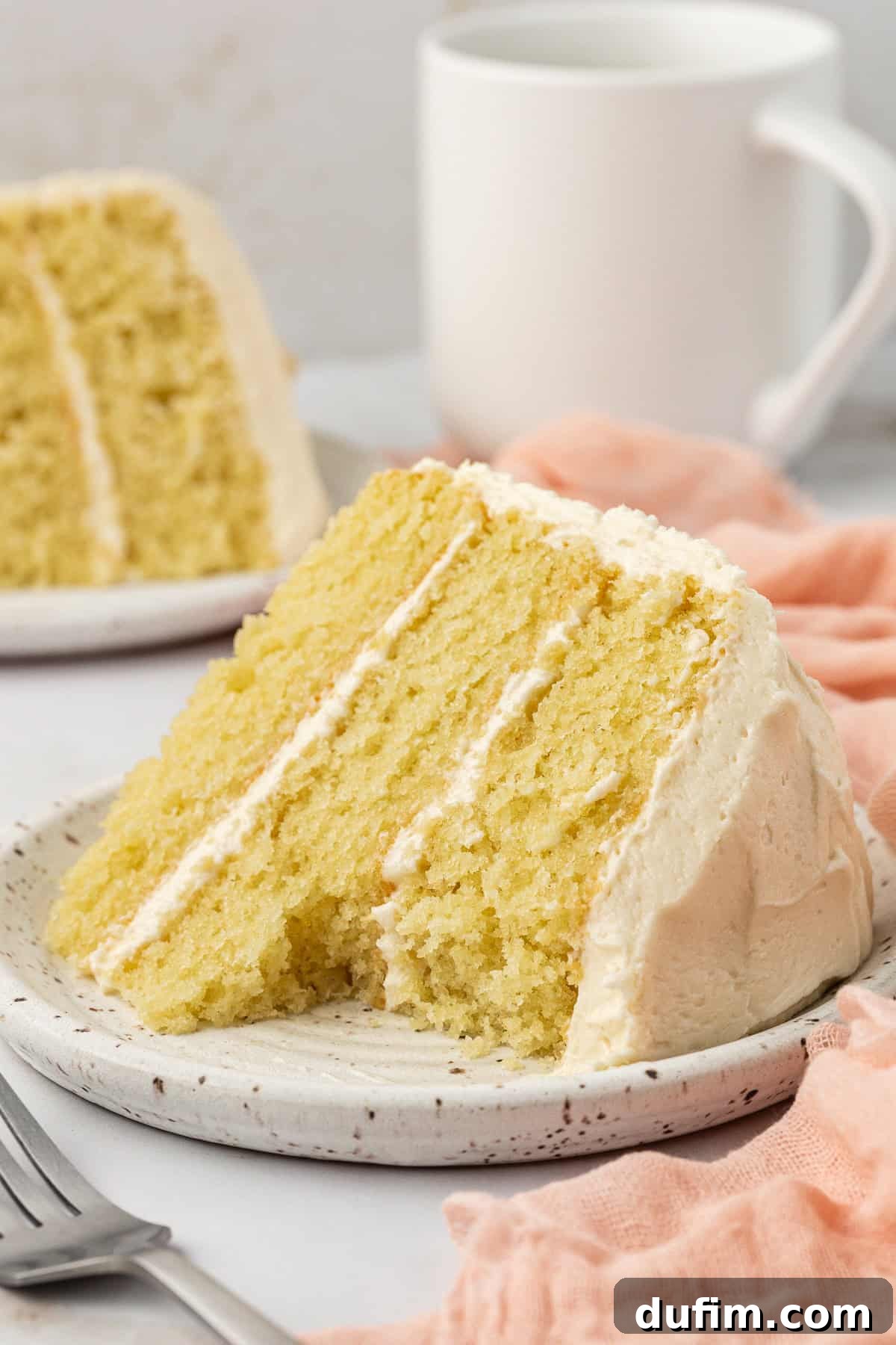 A tantalizing slice of vanilla cake with a bite missing, resting on a white speckled plate. A fork is nearby, and another slice of cake is blurred in the background, set on a pink cloth.