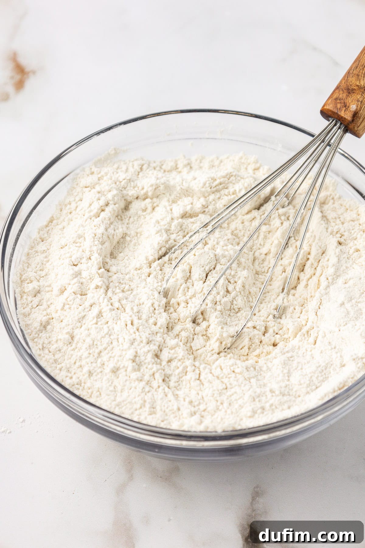 The dry ingredients for vanilla cake being whisked together in a clear glass bowl, ensuring they are thoroughly combined.