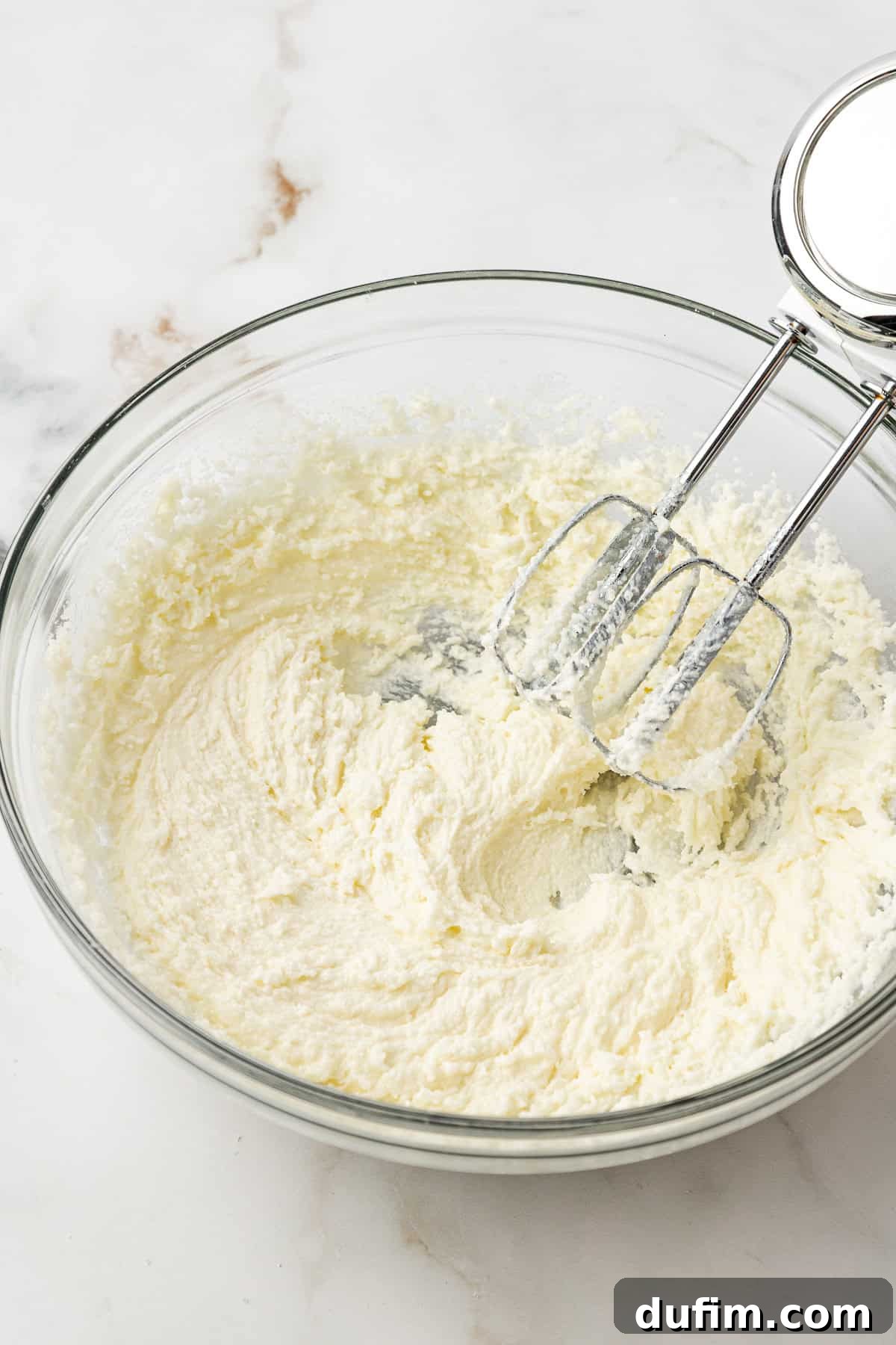 The butter, sugar, and oil mixture, after being beaten with an electric mixer, appearing light and fluffy in a clear glass bowl.