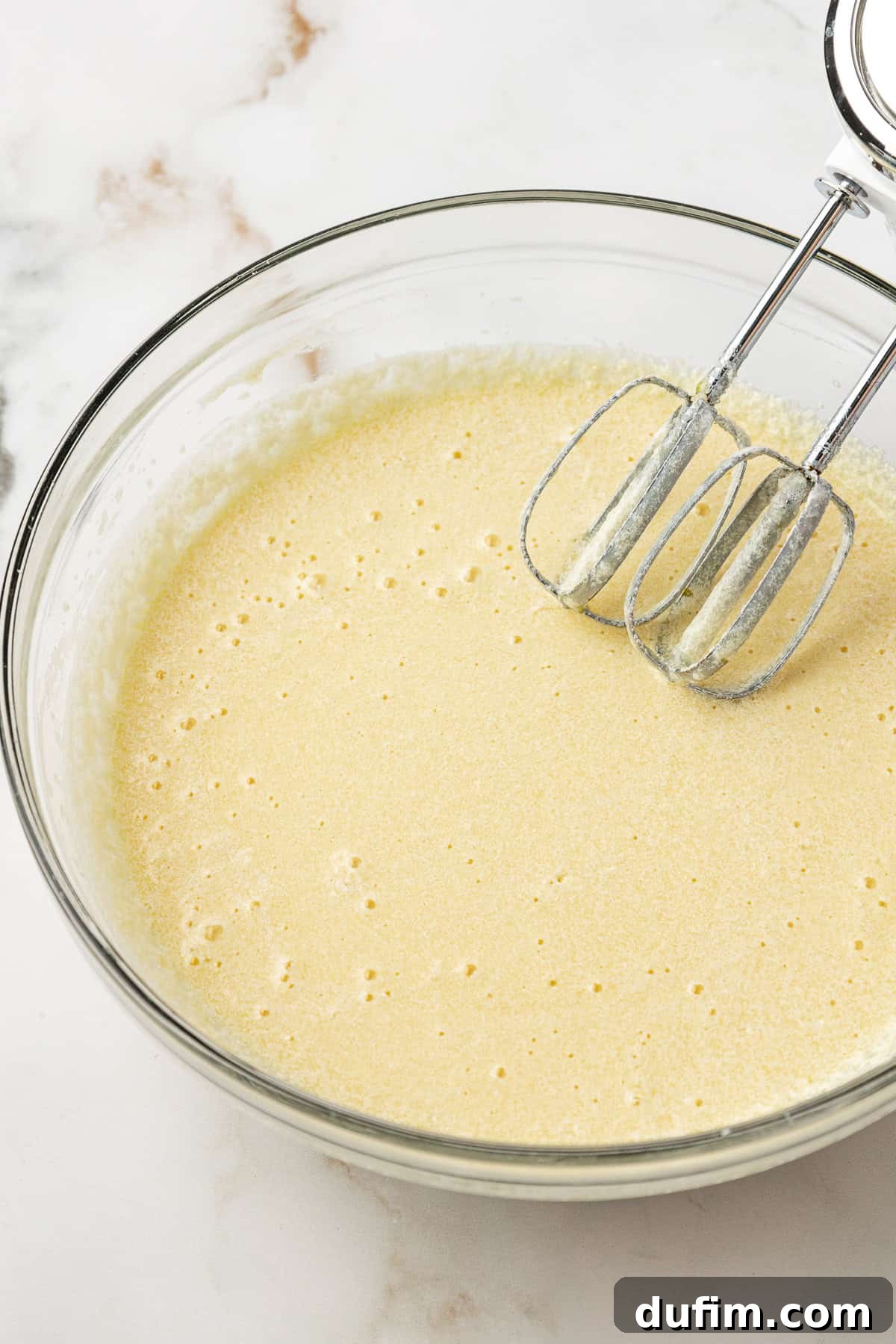 All the wet ingredients for the vanilla cake thoroughly mixed in a clear glass bowl with an electric mixer, forming a smooth base.