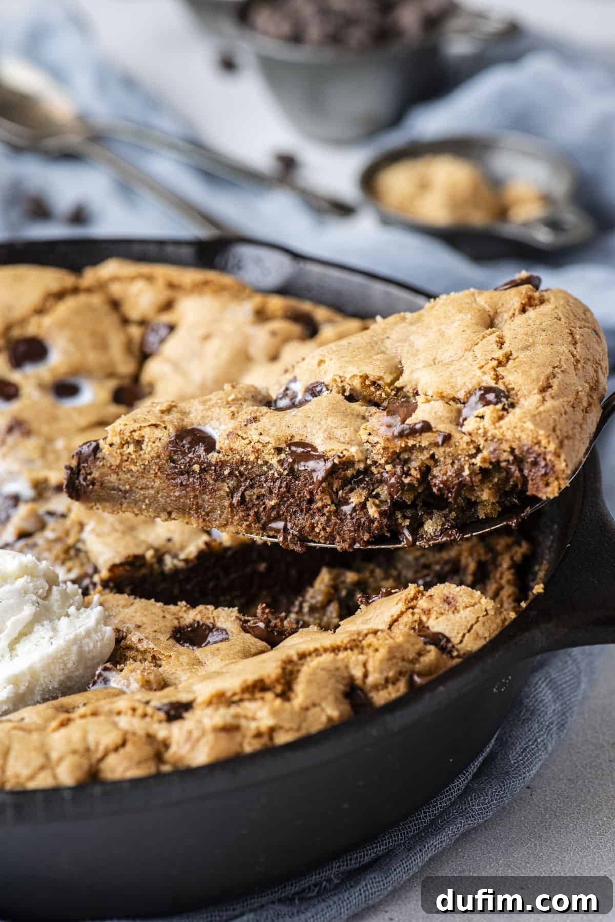 A slice of pizookie being carefully lifted from a rustic cast iron skillet, resting on a blue linen cloth. Scattered chocolate chips and spoons in the background hint at the deliciousness awaiting.
