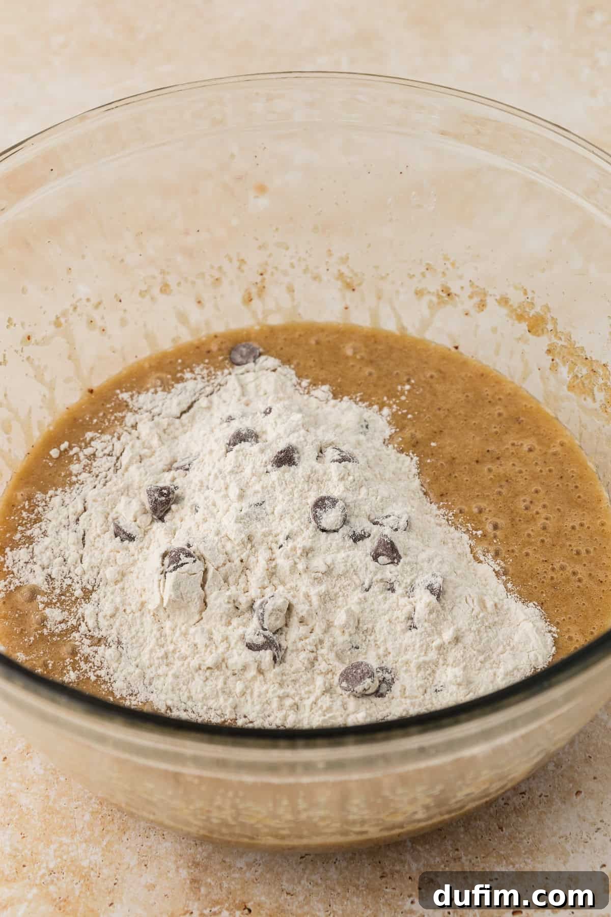 A clear glass bowl holds the wet ingredients for pizookie, with the dry ingredients neatly piled on top, poised for gentle mixing.