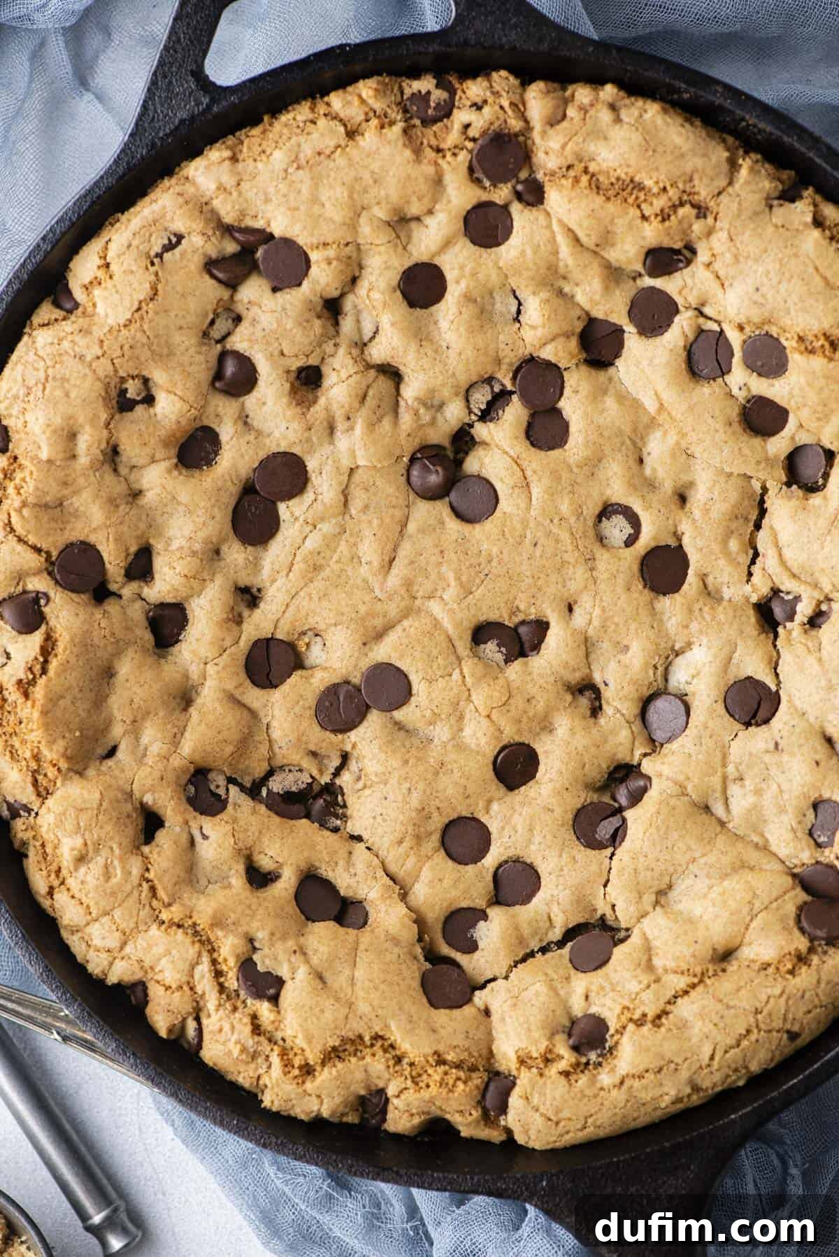An inviting overhead view of a freshly baked pizookie still in its cast iron skillet, resting on a soft blue linen cloth, its golden-brown edges promising deliciousness.