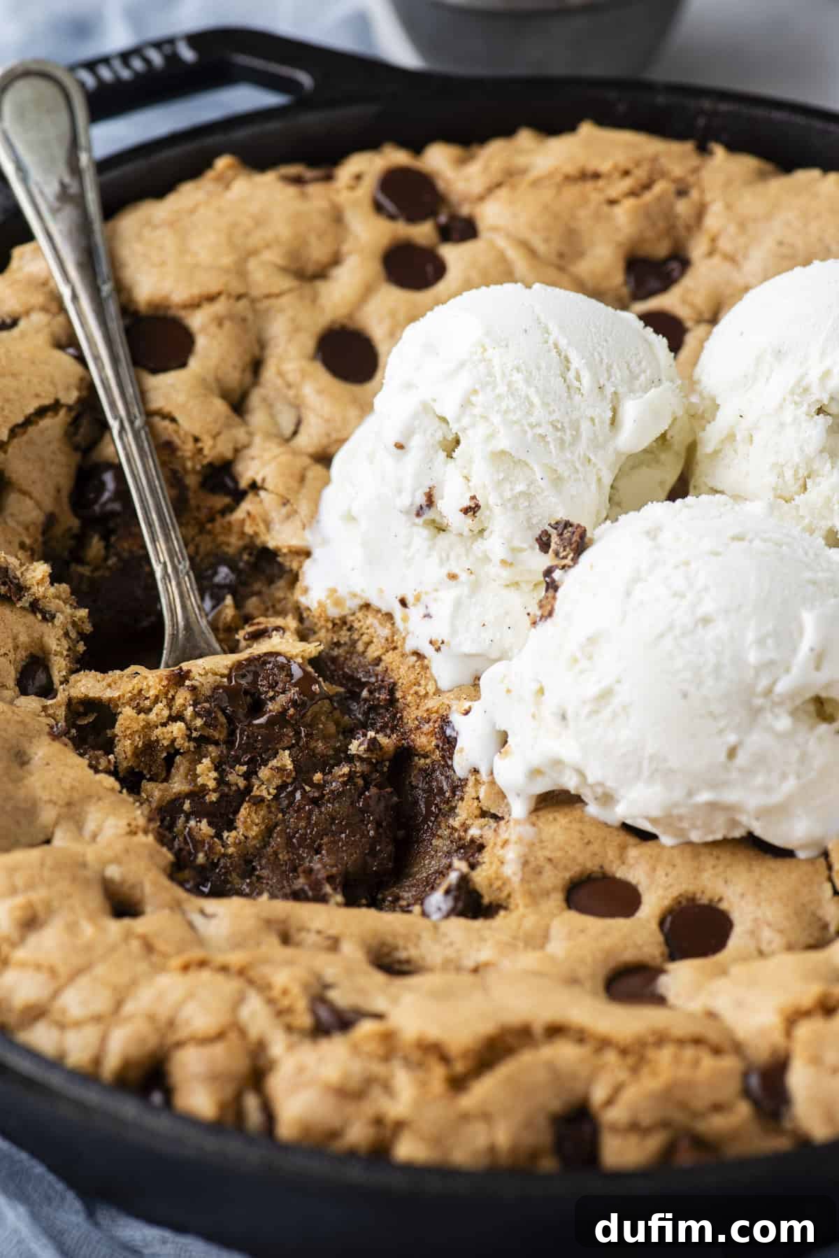 A beautifully baked pizookie nestled in a cast iron skillet, with a spoon plunged into its gooey center. Three generous scoops of vanilla ice cream sit atop the cookie, with inviting bites already taken, creating a tempting tableau.