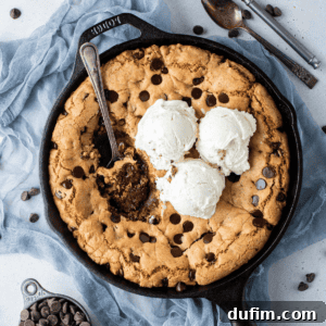 overhead view of a cast iron skillet full of a giant chocolate chip cookie (pizookie) and three scoop of vanilla ice cream, with a spoon in the middle and bites missing out, sitting on top of a blue cloth surrounded by a cup of chocolate chips, chocolate chips scattered around and two spoons