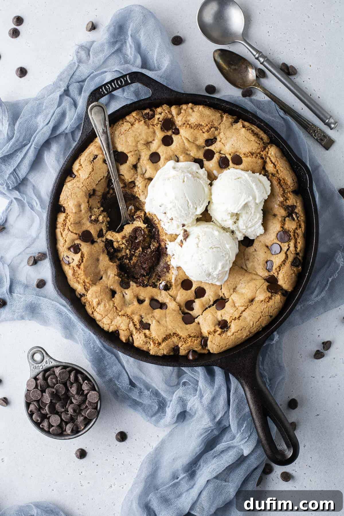 An inviting overhead shot of a cast iron skillet overflowing with a giant chocolate chip pizookie, topped with three generous scoops of creamy vanilla ice cream. A spoon is strategically placed in the center, with delightful bites already taken. The skillet rests on a blue cloth, surrounded by a cup of chocolate chips, scattered chips, and two additional spoons.