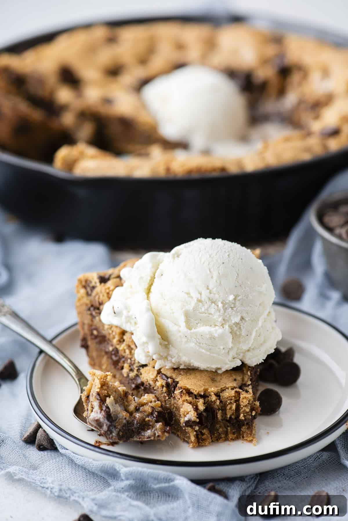A perfectly portioned slice of pizookie on a small white plate, crowned with a scoop of melting vanilla ice cream. A spoon with a bite of pizookie sits beside it, while the full cast iron skillet of pizookie awaits in the softly blurred background, surrounded by scattered chocolate chips.