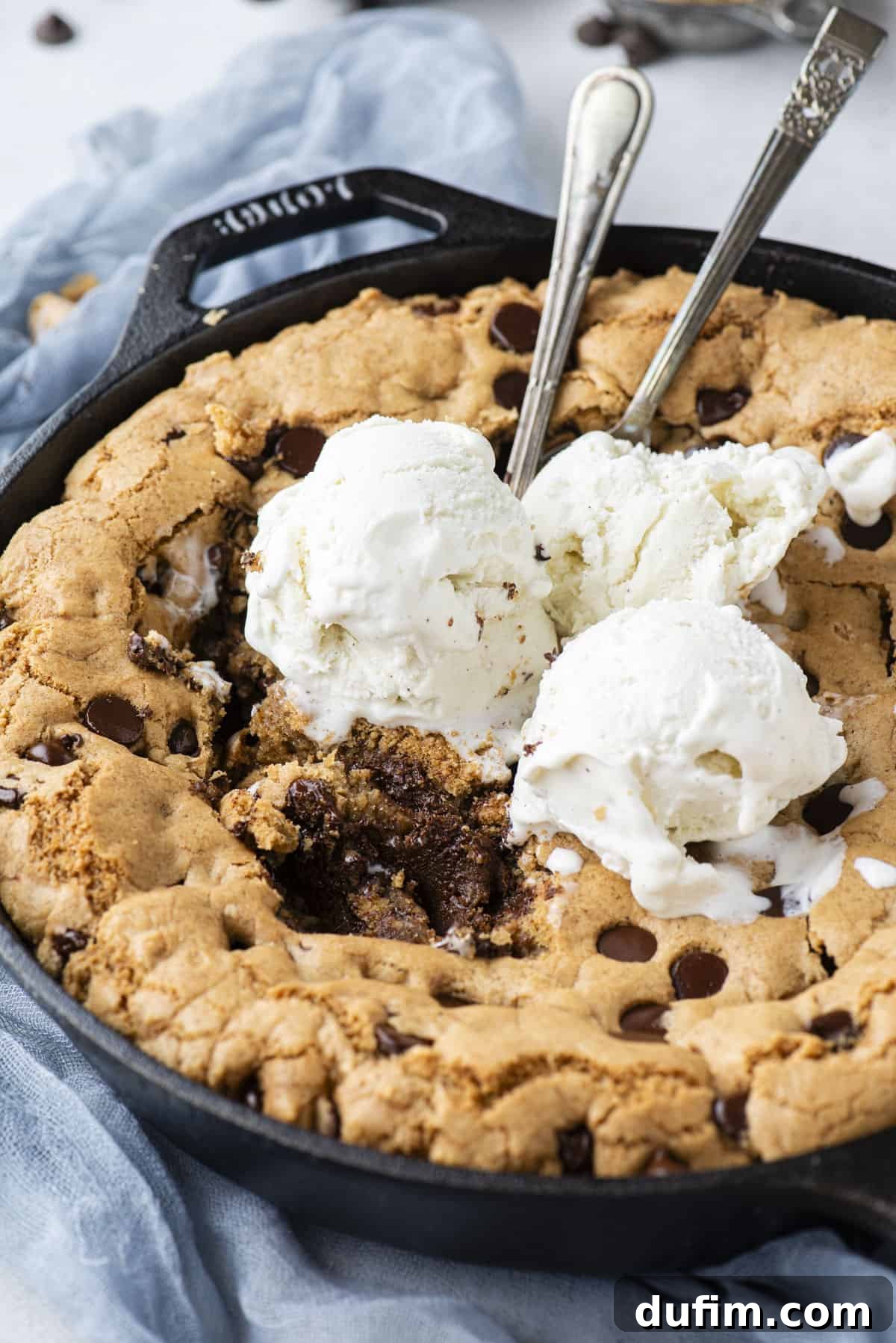 A rustic cast iron skillet filled with a freshly baked pizookie, artfully topped with three generous scoops of vanilla ice cream. Bites are already missing from the center, and two spoons are eagerly stuck into the dessert, all resting on a soft blue linen cloth.