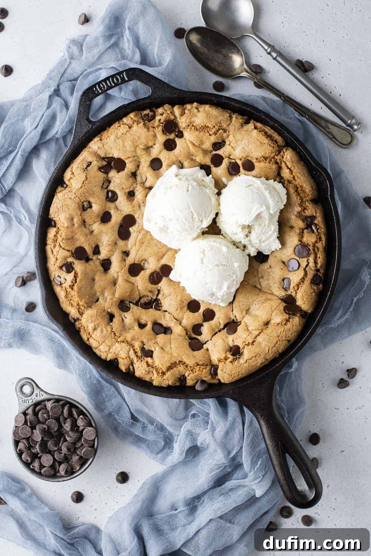 An inviting overhead view of a cast iron skillet brimming with a baked pizookie, adorned with three scoops of vanilla ice cream. The skillet sits on a blue cloth, surrounded by scattered chocolate chips, a small cup of additional chocolate chips, and two awaiting spoons.