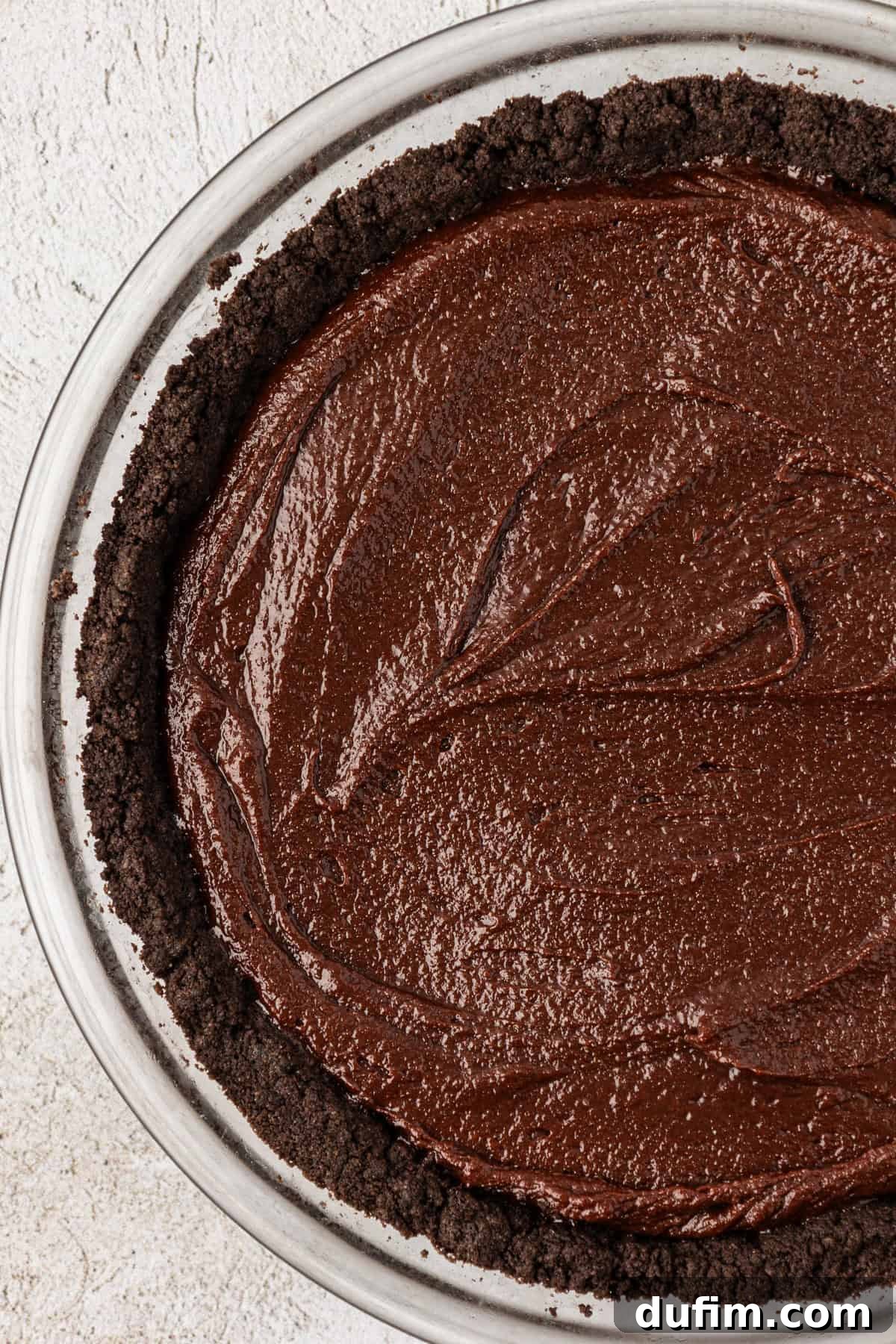 a clear glass pie dish with an oreo crust layer and a chocolate brownie layer that hasn't been cooked yet