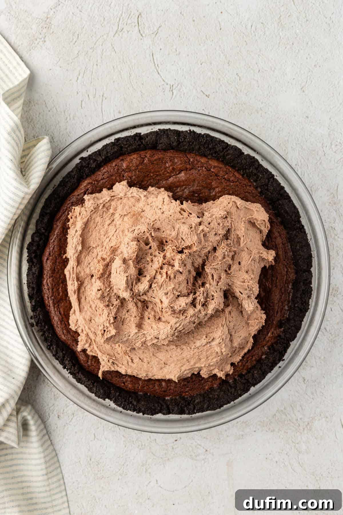 a glass pie dish with oreo crust and a baked brownie layer, with a mound of chocolate mousse on top that hasn't been spread yet