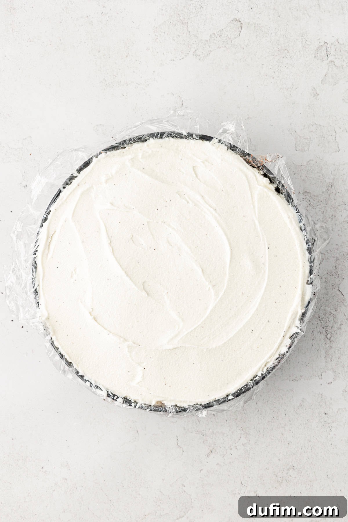 An overhead shot of a partially completed ice cream cake, with the top layer covered in plastic wrap for freezing.