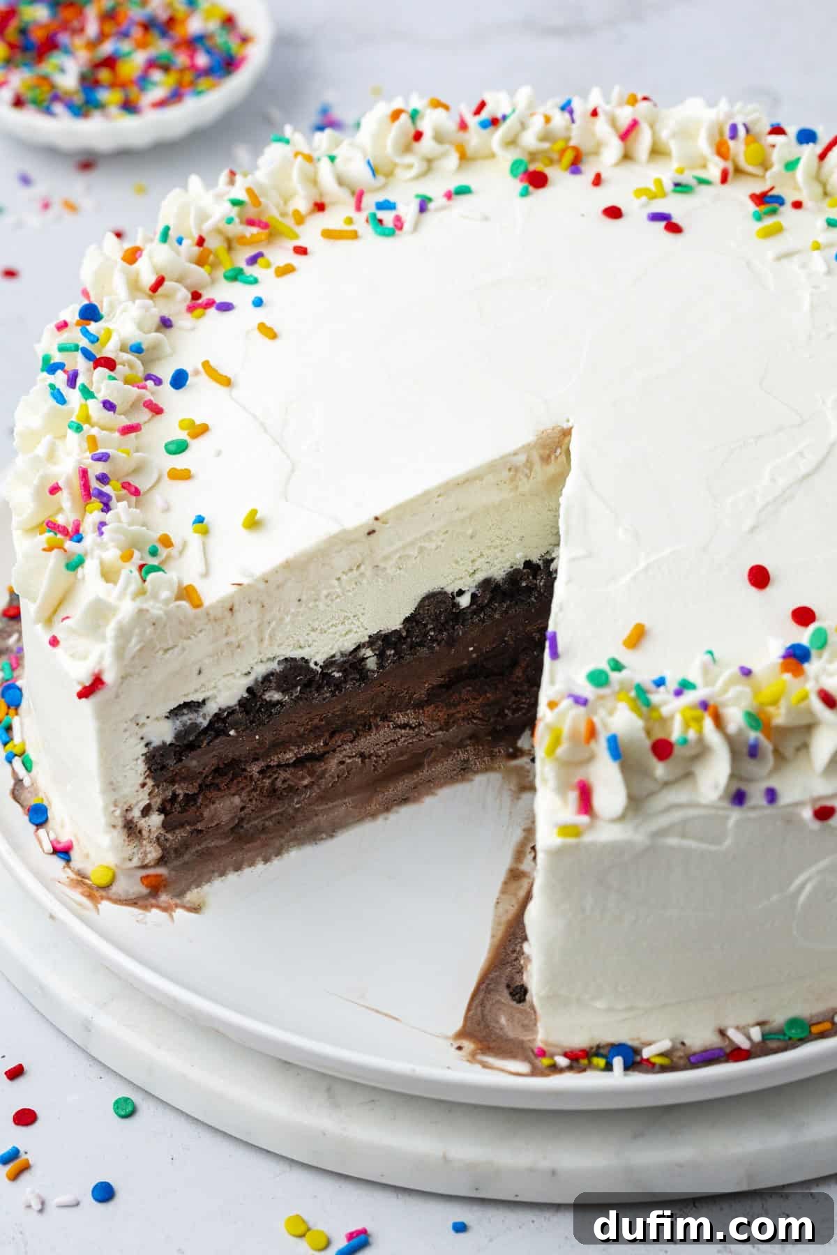 A vibrant ice cream cake topped with a generous amount of rainbow sprinkles, with one slice thoughtfully removed, and more sprinkles scattered artfully around a small bowl in the background.