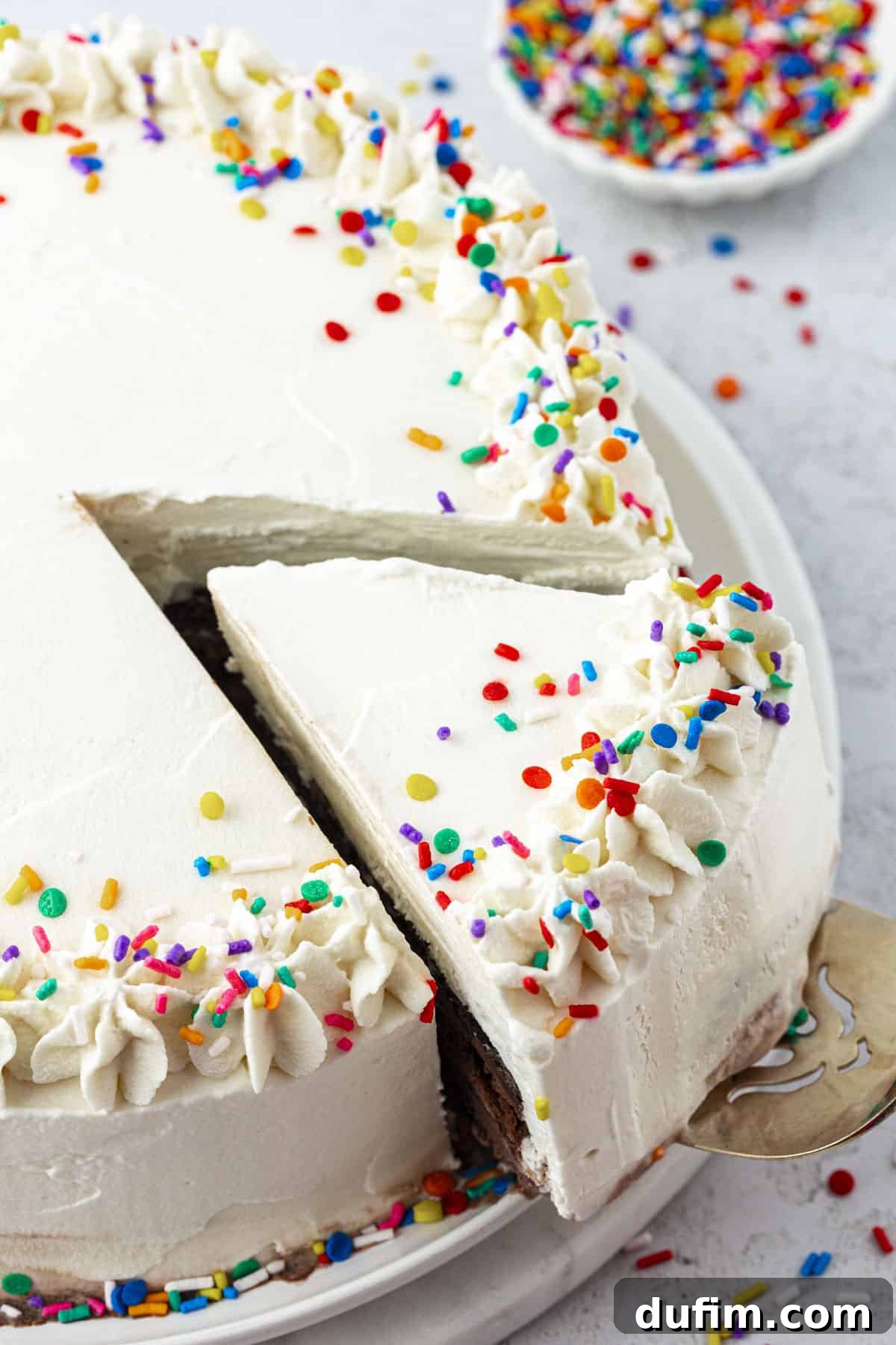 An inviting overhead view of a rainbow-sprinkled ice cream cake, with one slice lifted, showcasing its layers, and a small bowl of sprinkles nearby.