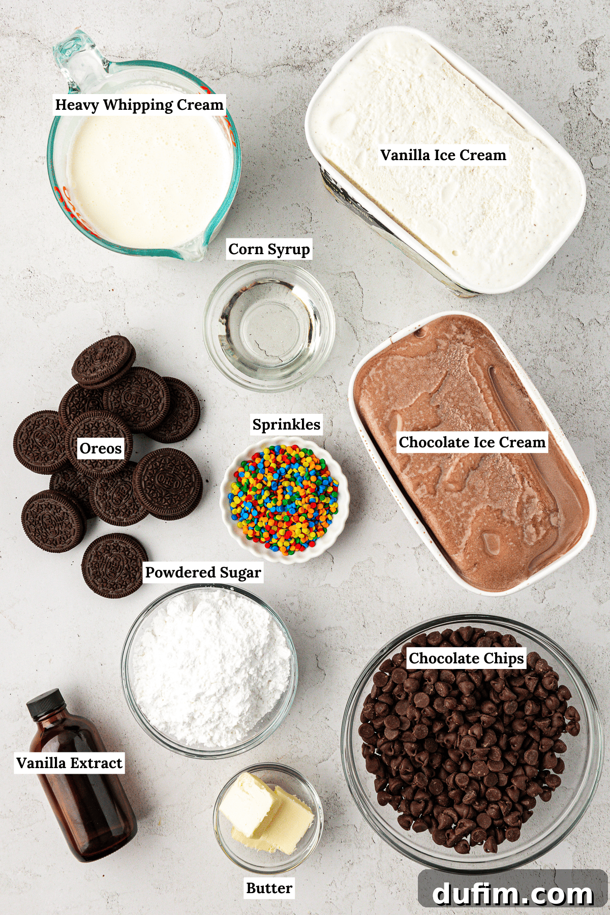 Overhead view of all the essential ingredients laid out for making an ice cream cake: heavy whipping cream, vanilla ice cream, corn syrup, Oreos, colorful sprinkles, chocolate ice cream, powdered sugar, vanilla extract, butter, and chocolate chips.