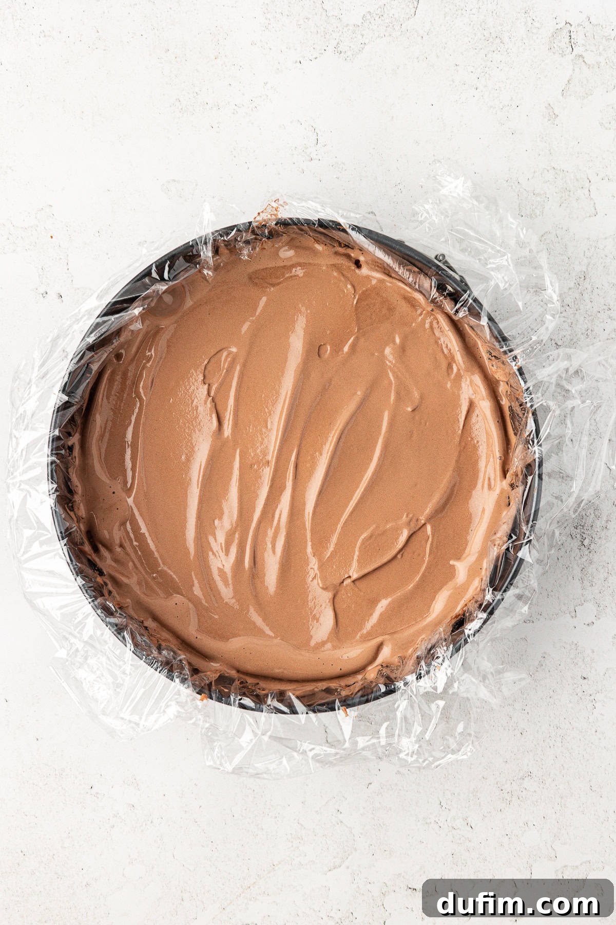 The first layer of chocolate ice cream spread evenly within a plastic-wrapped springform pan, awaiting the next delicious layer.