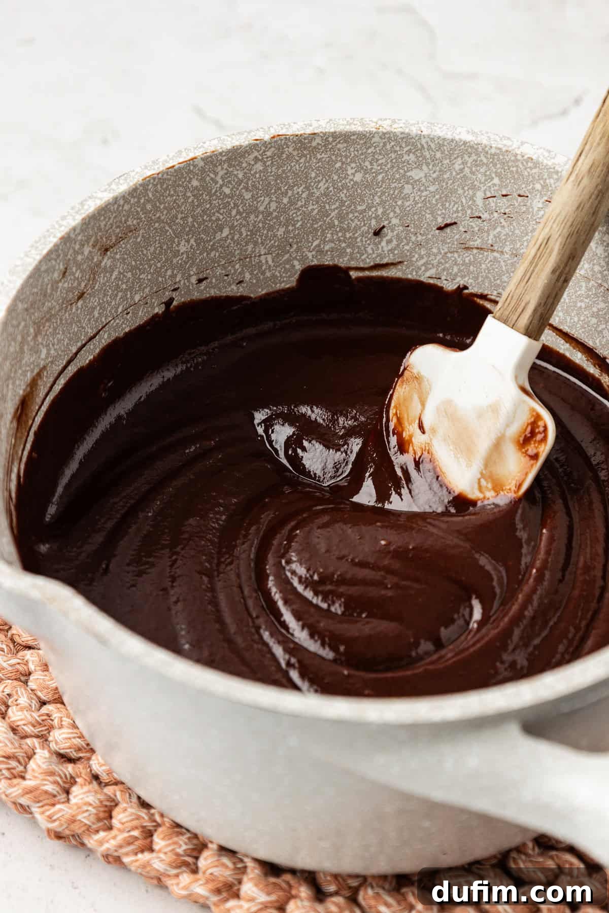 Rich chocolate fudge ganache being stirred with a white spatula in a small saucepan, showcasing its smooth, luscious texture.